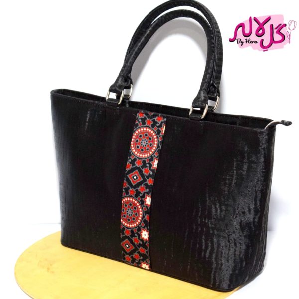 Gleaming Black - Faux Leather Hand Bag A shiny beautiful handbag with a traditional twist. Featuring a simple & popular silhouette, this bag is perfect to carry everything you need to have with you when on the go! Made out of faux leather with traditional printed fabric incorporated to make it stand out and give it a desi touch! Colour: Black